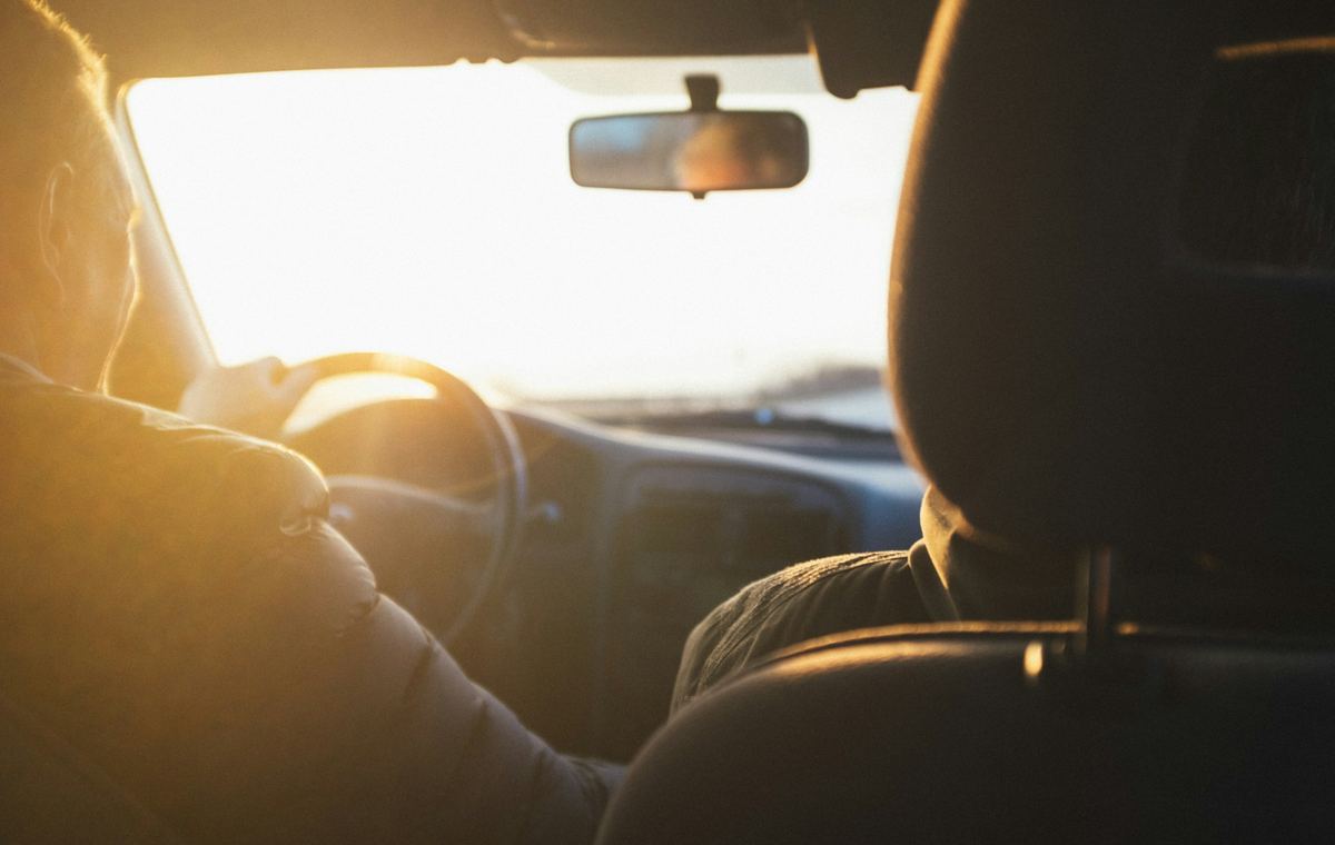 Iemand achter het stuur van een wagen tijdens een rit in het avondlicht, gezien vanaf de achterbank
